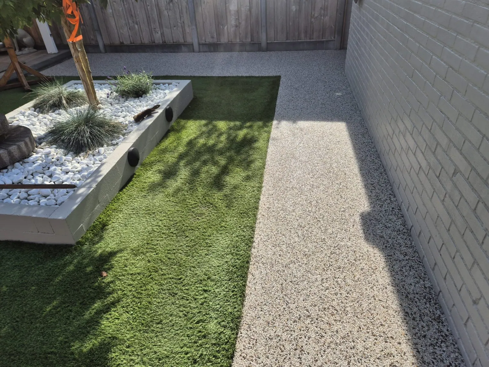 Smal tuinpad van harsgebonden grind langs kunstgras en een witte plantenbak.