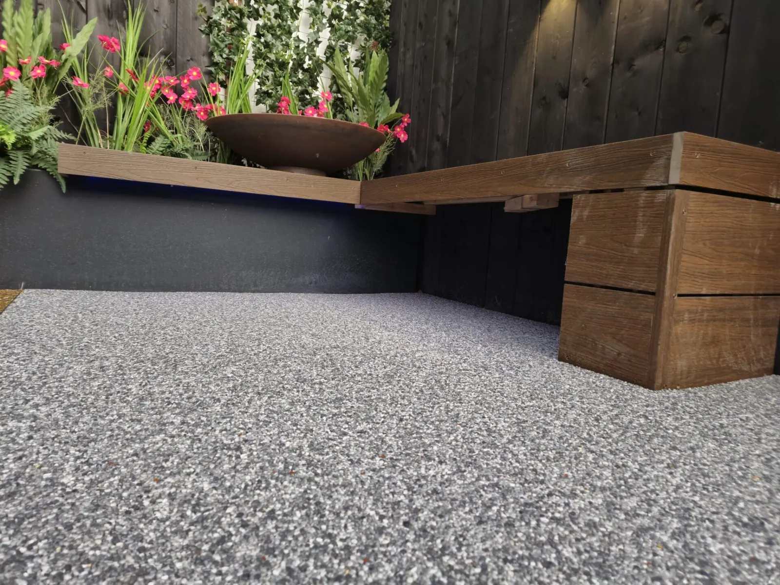 Vloer van harsgebonden grind bij een houten zitbank en plantenbak met bloemen, tegen een donkere schutting.