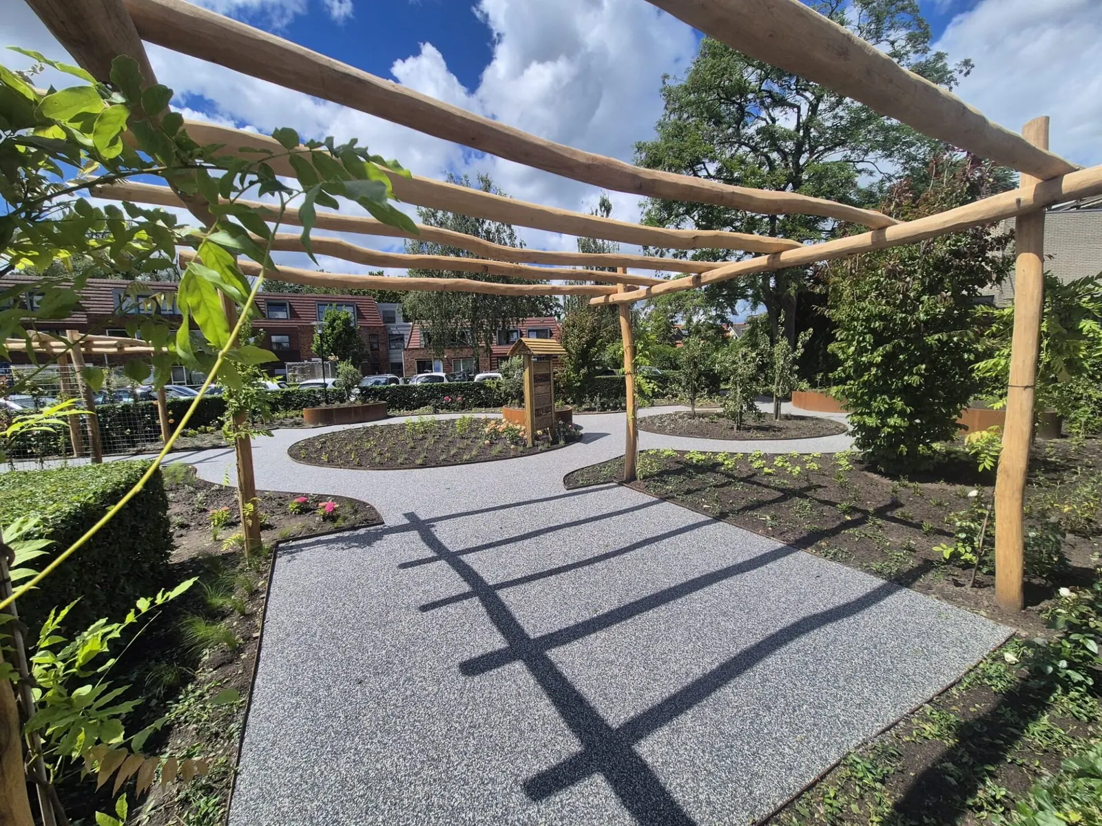 Grindpad onder een houten pergola met schaduwlijnen, tussen borders met jonge beplanting.