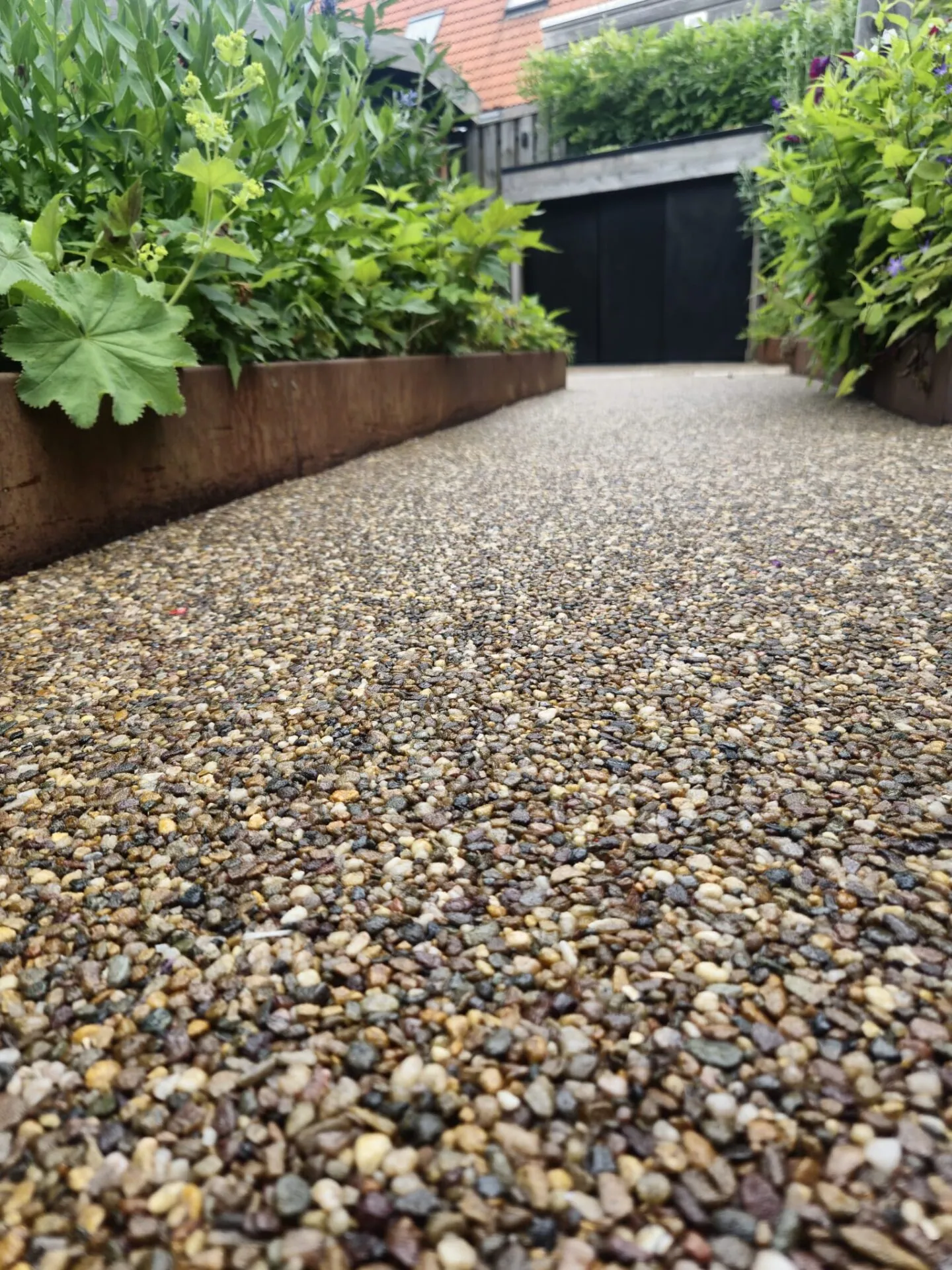 Grindpad tussen cortenstalen plantenbakken met weelderige beplanting, richting een zwarte schutting.