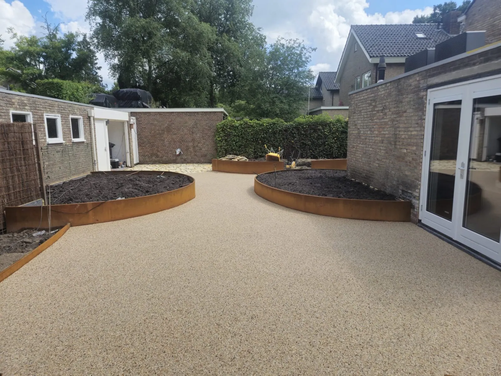 Groot beige terras van harsgebonden grind met ronde cortenstalen plantenbakken
