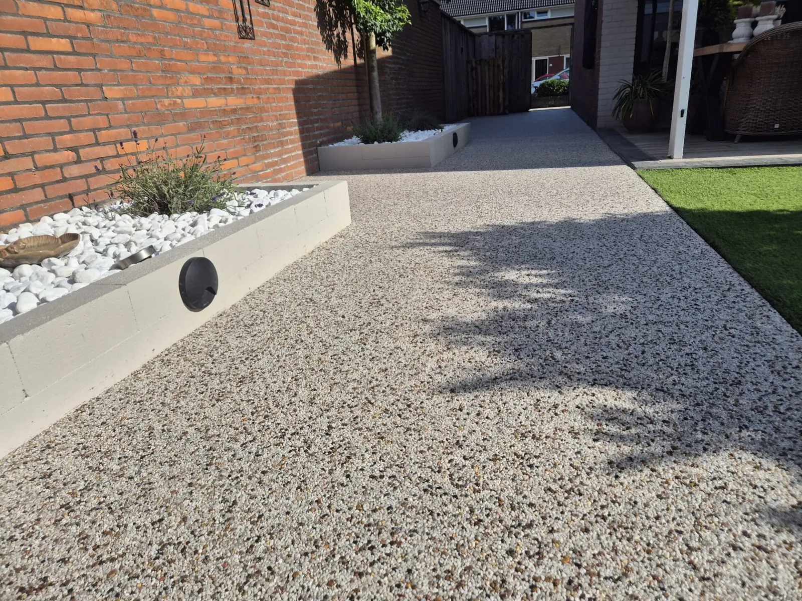 Pad van harsgebonden grind naast de woning, met een witte verhoogde plantenbak en boomschaduw.