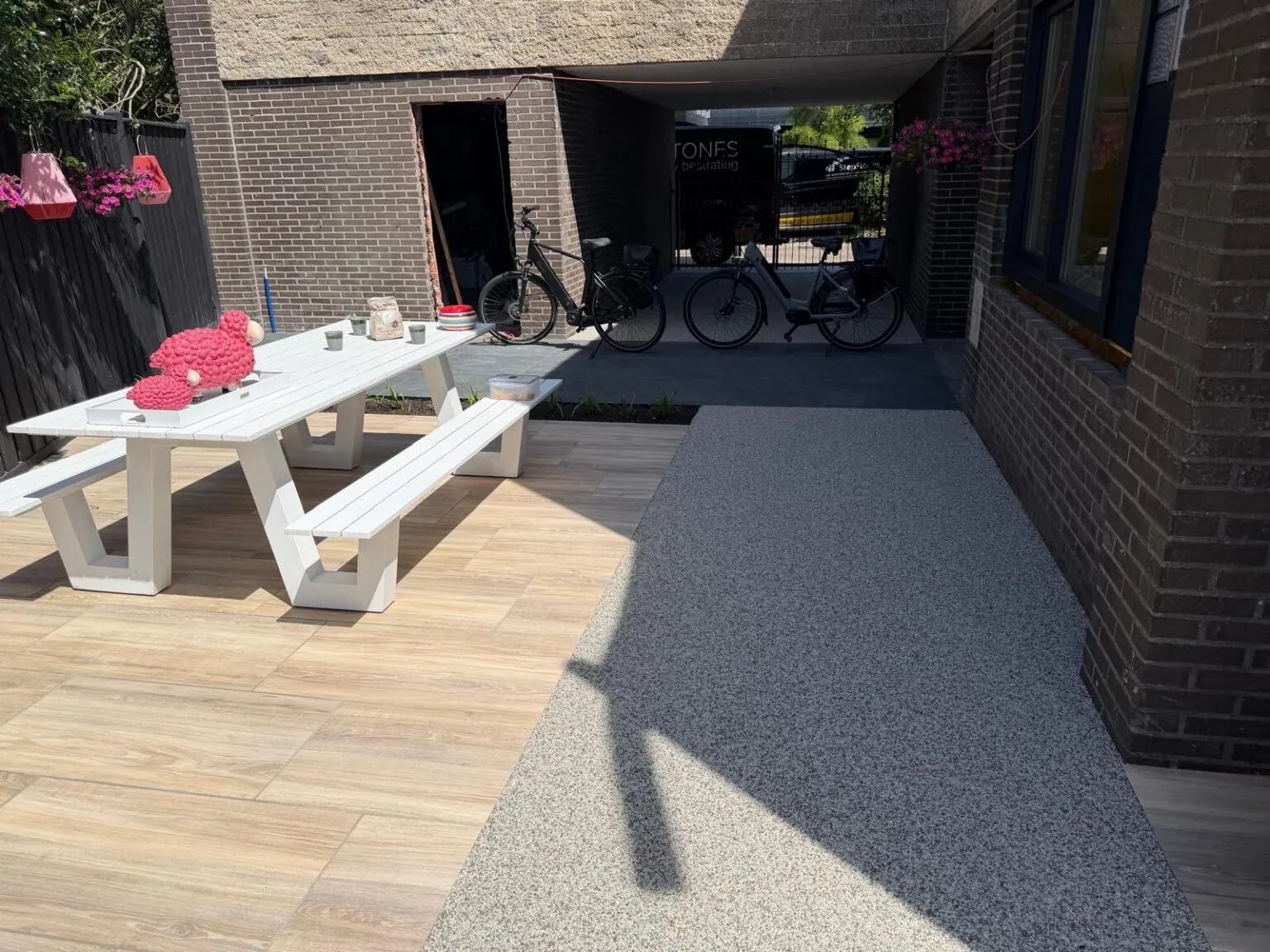 Vloer van harsgebonden grind naast keramische tegels in houtlook, met picknicktafel bij een overkapping.