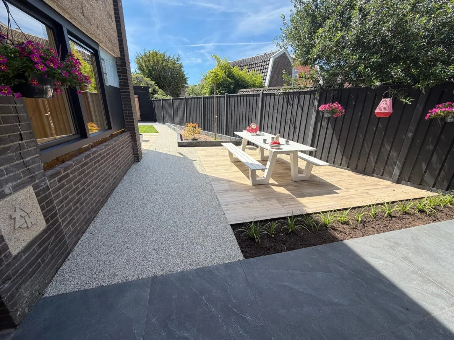 Grindpad langs de woning en een plantenborder naar een houten vlonder met picknicktafel.