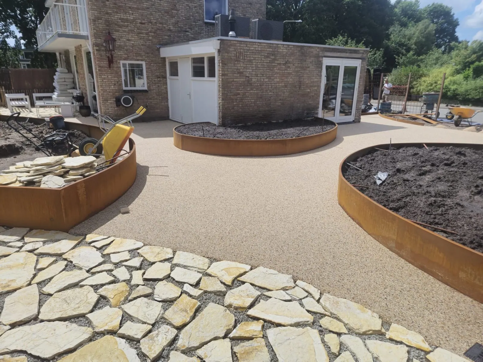 Terras van harsgebonden grind met ronde cortenstalen plantenbakken bij een tuinhuis en natuurstenen flagstones.
