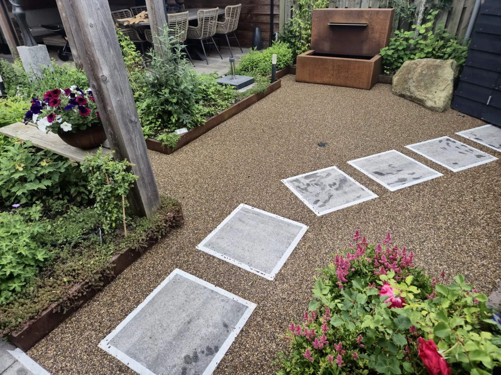 Grindterras met losse tegels als stapstenen, bloeiende planten en een zithoek op de achtergrond.