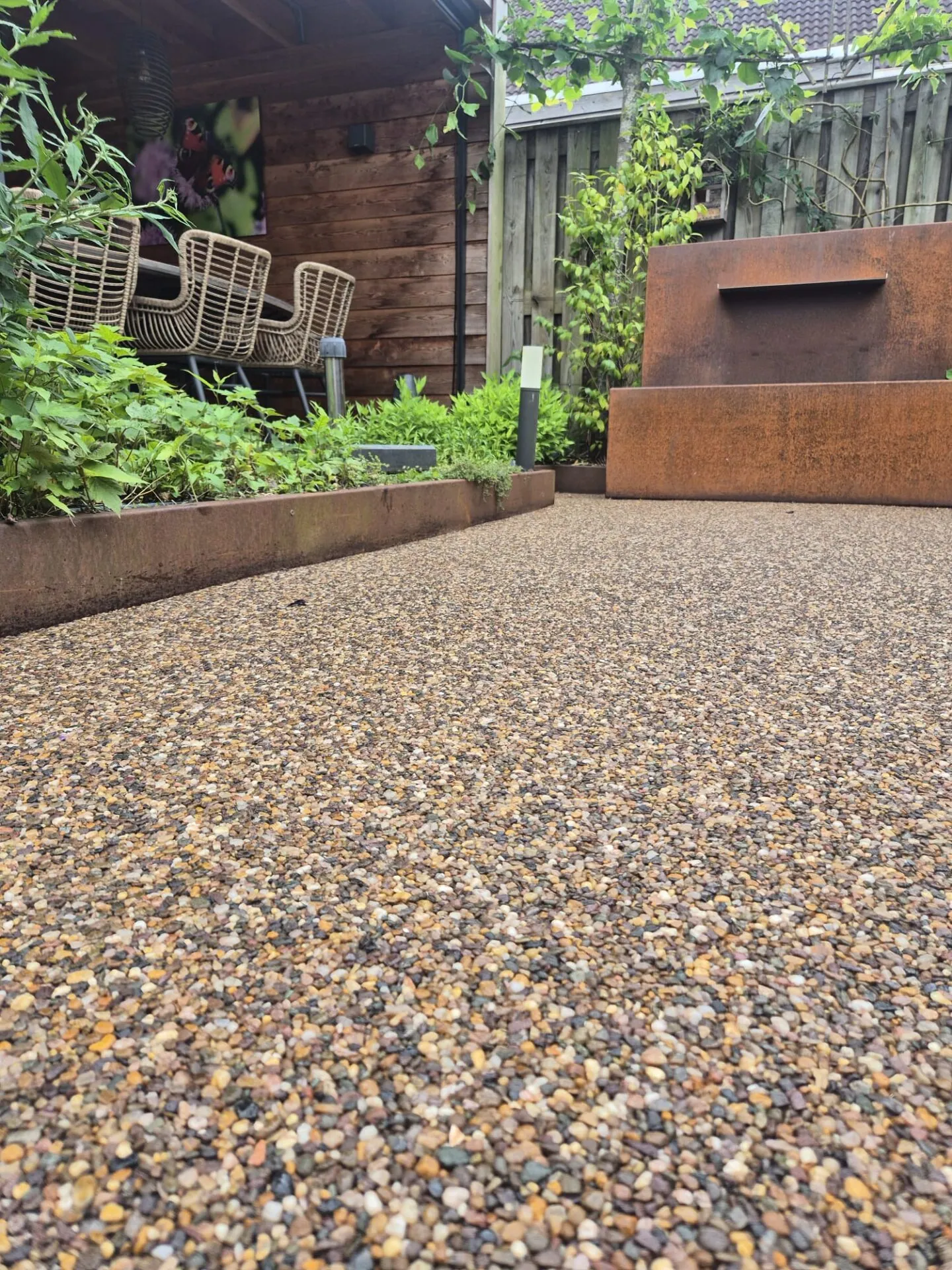 Terras van harsgebonden grind met cortenstalen plantenbakken, een zithoek en een waterornament.